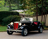 AUT 21 RK0286 02
1952 Black MG TD Convertible 3/4 Front View On Pavement Headlights On By Trees And Bushes