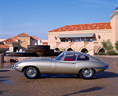 AUT 12 RK0013 08
1961 Jaguar XKE Coupe E-Type Gold Profile On Pavement By Building And Water Fountain Blue Sky
