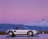 POR 04 RK0201 04
1998 Porsche Boxster Convertible Silver Low Profile On Wet Pavement Tall Grass & Mts In Background Headlights On