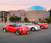POR 07 RK0121 01
1957, 1989, 1994 Porsche Speedster Convertibles Silver And Red 3/4 Front View On Pavement By Building